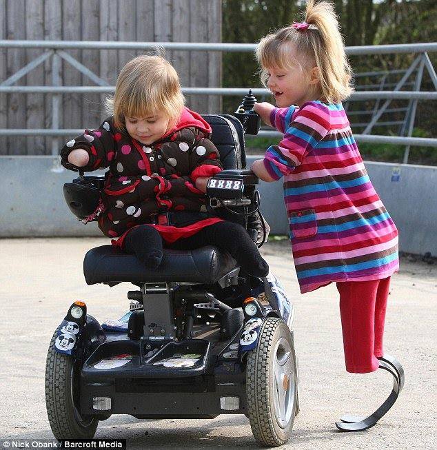 Une fillettes sur des protheses en lames qui surveille une autre plus petite dans son fauteuil electrique
